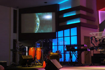 close up of video monitor screen with video side view of the earth on far side wall, percussion and 		synthesizer instruments on stage with translucent panel with vibrant blue LED behind it, International 	Tabernacle, West Palm Beach, FL, USA