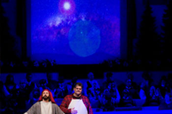 evening scene with video screen of a space nebula with hues of blues, whites and reds, blue is cast over the singers on stage and a spot light on two men acting - Journey to the Manger Musical - South Orlando Baptist Church, Orlando, FL, USA
