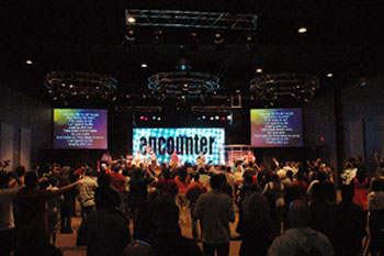 congregation with their hands in the air with a band performing on stage illuminated by white and red LEDs, screens with lyrics to a song and a LED video wall that says encounter  in the Word of Life Center - Shreveport, Louisiana, USA
