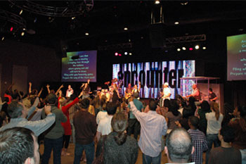 congregation with their hands in the air with a band performing on stage illuminated by white and red LEDs, screens with lyrics to a song and a LED video wall that says encounter  in the Word of Life Center - Shreveport, Louisiana, USA