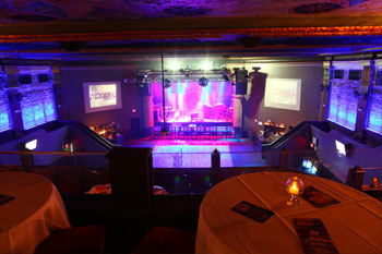 upper mezzanine dining area covered tables LED candles on top and bench chairs with a view of the floor and stage. Walls of theatre vibrant blues stage magenta and white screens on both sides of stage say The Beacham inside The Beacham - Orlando, Florida, USA