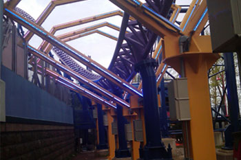 blue rollercoaster tracks going through yellow diamond shaped metal structures with mounted lit LED Pulsar Chromastrip fixtures with vibrant blue color and  gray juncture boxes mounted at the base of them on the Bizarro Coaster Thrill Ride at night, Six Flags New England - Agawam, Massachusetts, USA