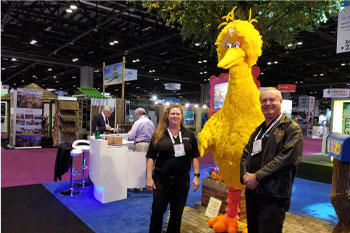 techni-lux in-house lighting designer tony hansen and field installation consultant lisa hansen posing with Sesame Street character Big Bird with his bright yellow feathers and beak and bright orange feet at Sally Dark Rides booth IAPPA for Seasame Street: Street Mission dark ride - PortAventura World, Salou, Spain