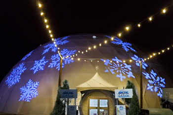 snowflake patterned gobos illuminated in white exterior Artic Igloo with strands of round white LED lights above on poles in front of entrance at Snowcat Ridge Alpine Snow Park - Dade City, Florida, USA