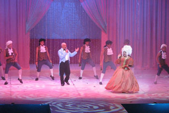 male dancers dressed in 18th century frocks with a man in a blue tuxedo and tails holding a microphone singing to a women dressed in an 18th century gown, backdrop red curtains and another red curtain drawn open spotlights on the performers and bright white geometric gobo patterns on the stage floor in the play Glamur, Sexo, Divas y Otras Mentiras - Teatro Nacional, Dominican Republic